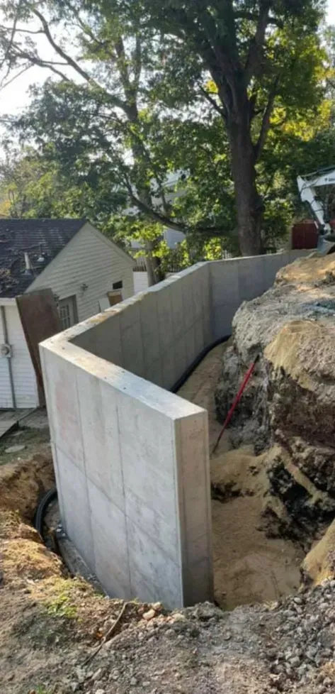Concrete retaining wall around a rocky slope, with trees and a house in the background.
