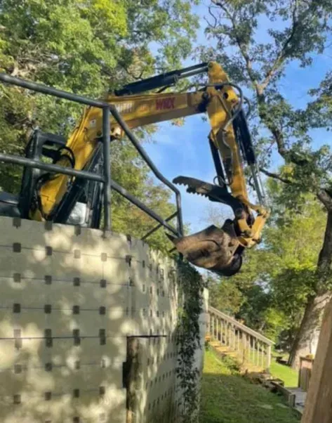 Yellow excavator lifting debris beside a stone wall in a wooded area.
