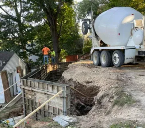 Construction crew pouring concrete into a basement foundation beside a cement truck on a sloped lot