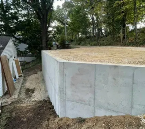Concrete foundation wall under construction beside a wooded yard and small house