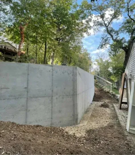 Concrete retaining wall beside a dirt path on a wooded hillside, with a house at right and stairs in back.