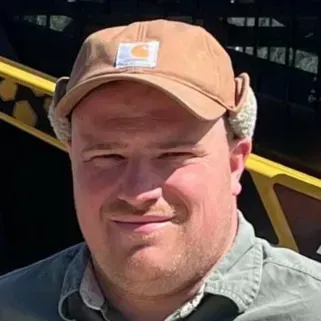 A person in a tan cap and green shirt stands outdoors in bright sunlight.