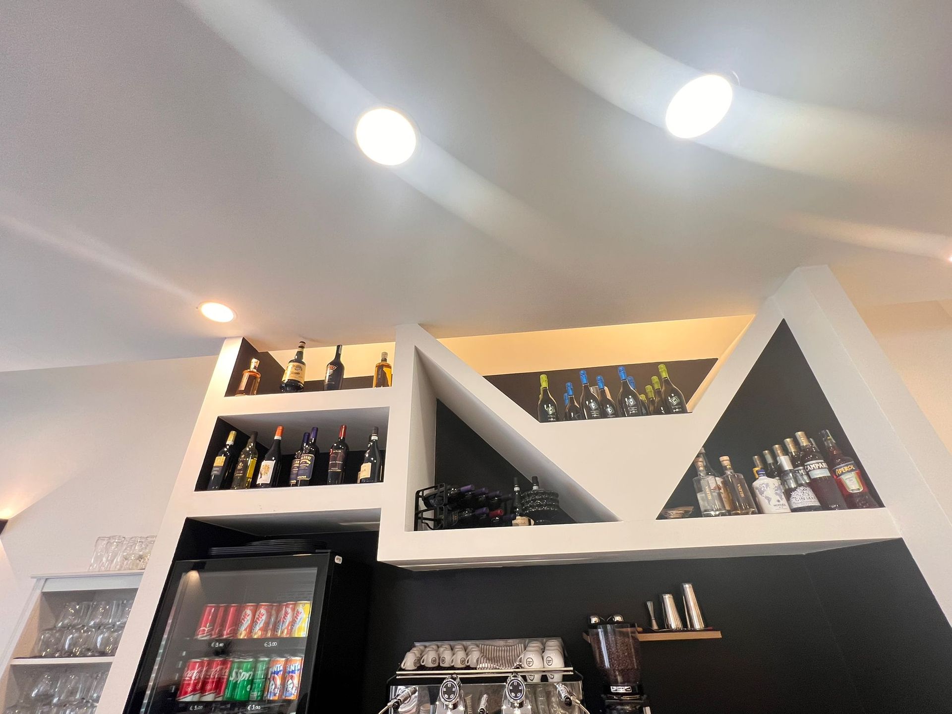 Interno del bar con bottiglie di liquore sugli scaffali, frigorifero nero e illuminazione dall'alto.