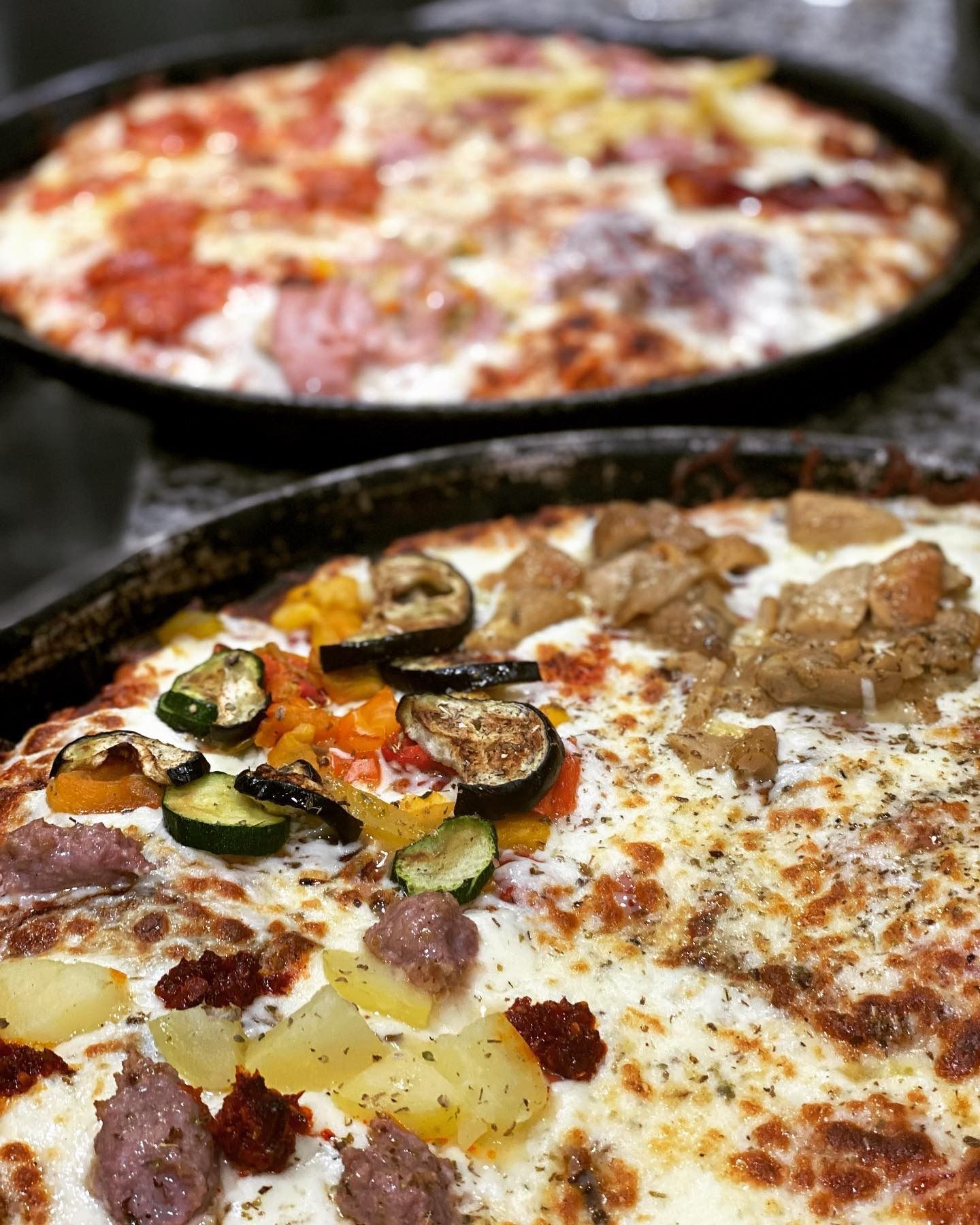 Due pizze in teglie scure, una con verdure e salsiccia, l'altra con salame piccante e altri condimenti.