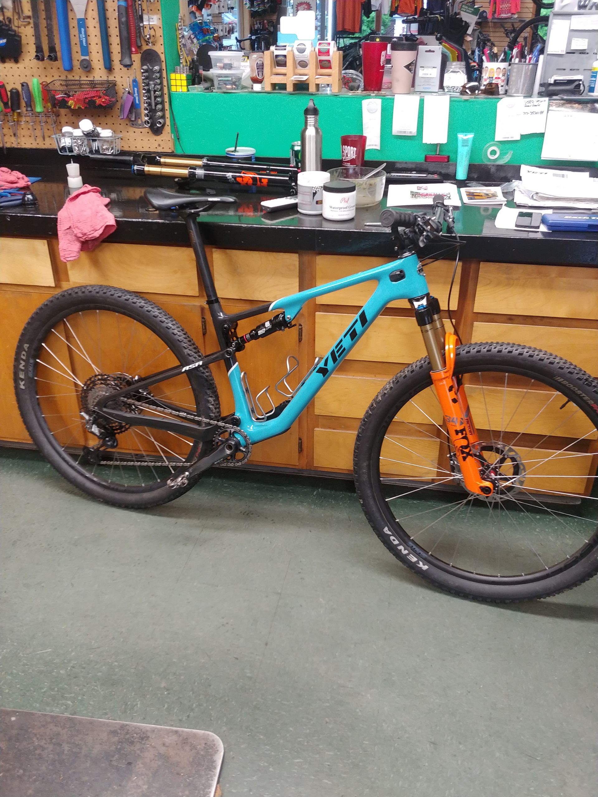 Blue-and-orange mountain bike displayed indoors on a shop floor in front of a workbench
