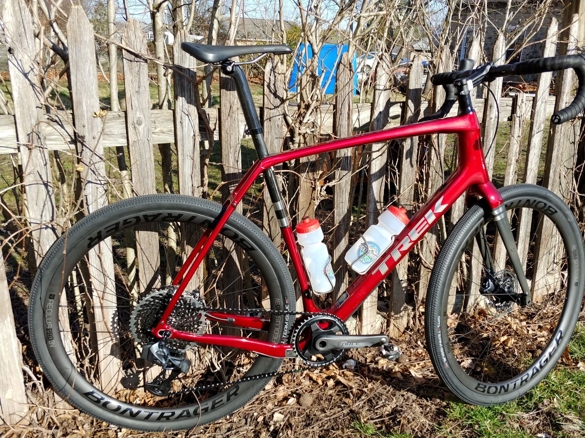 Red road bike leaning against a fence in a leaf-covered yard, with water bottles attached to the frame