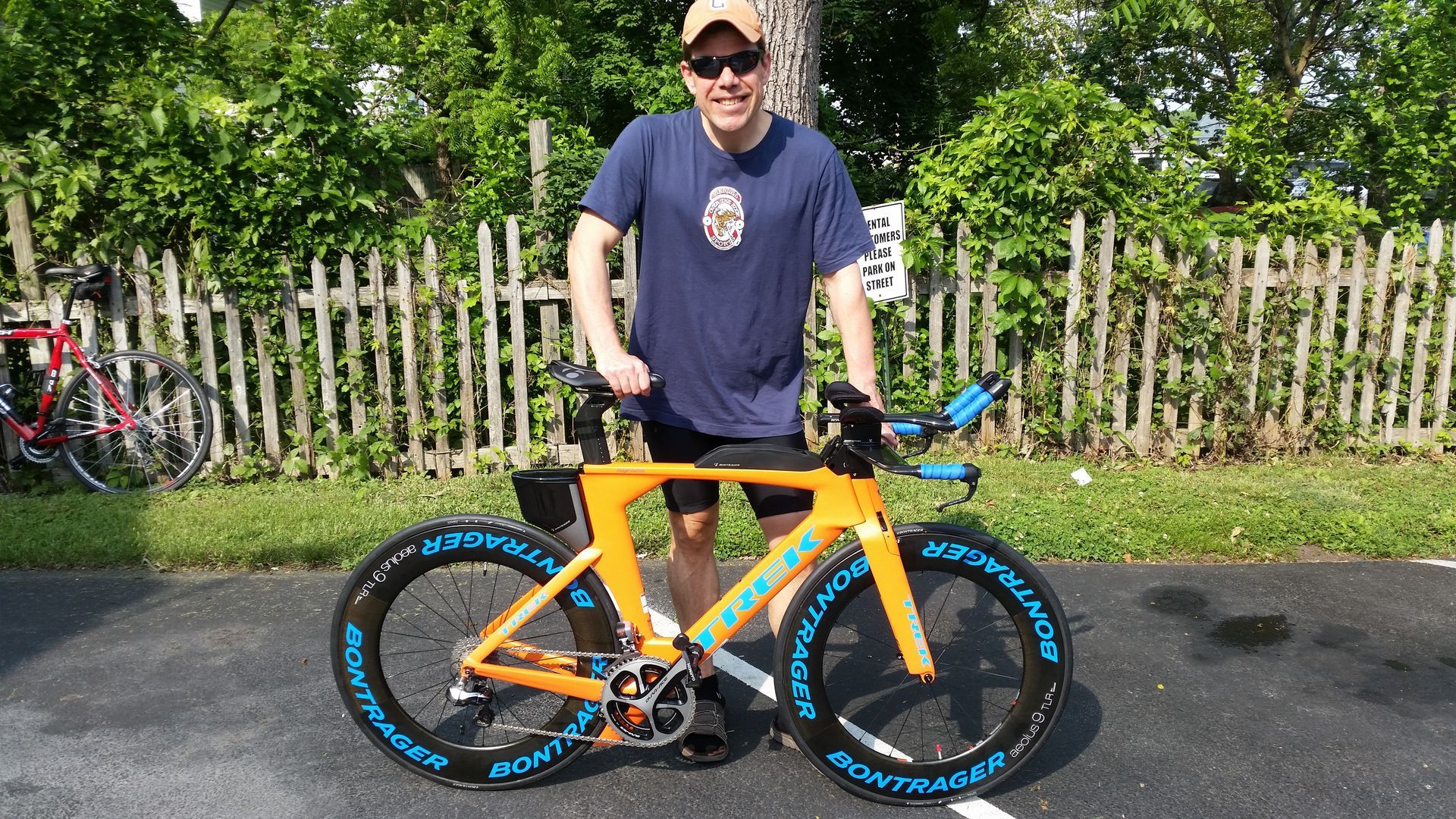 Person standing with an orange-and-blue bicycle on a road beside a fence and greenery.