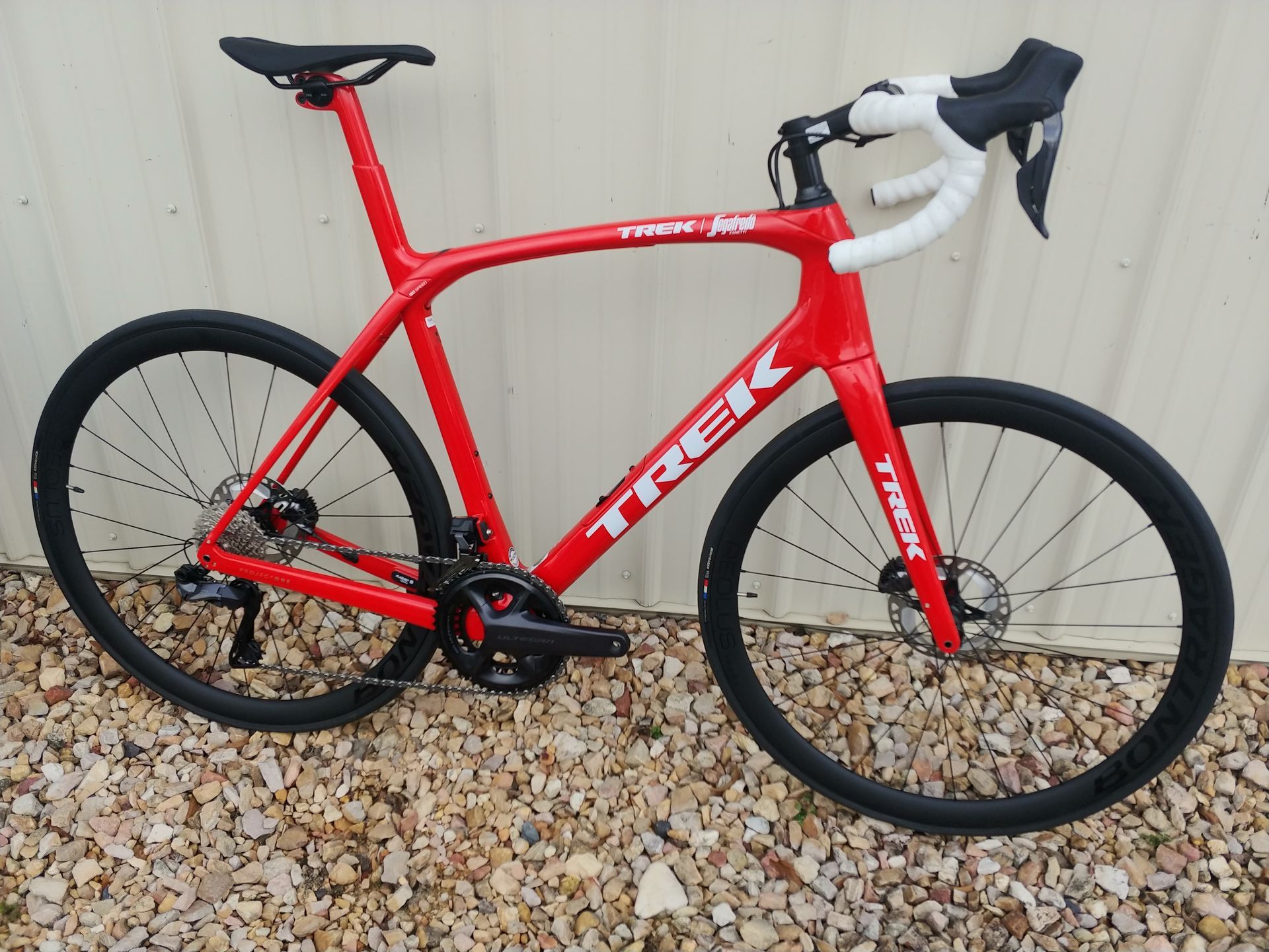 Red Trek road bike with white handlebars, leaning against a beige wall on gravel.