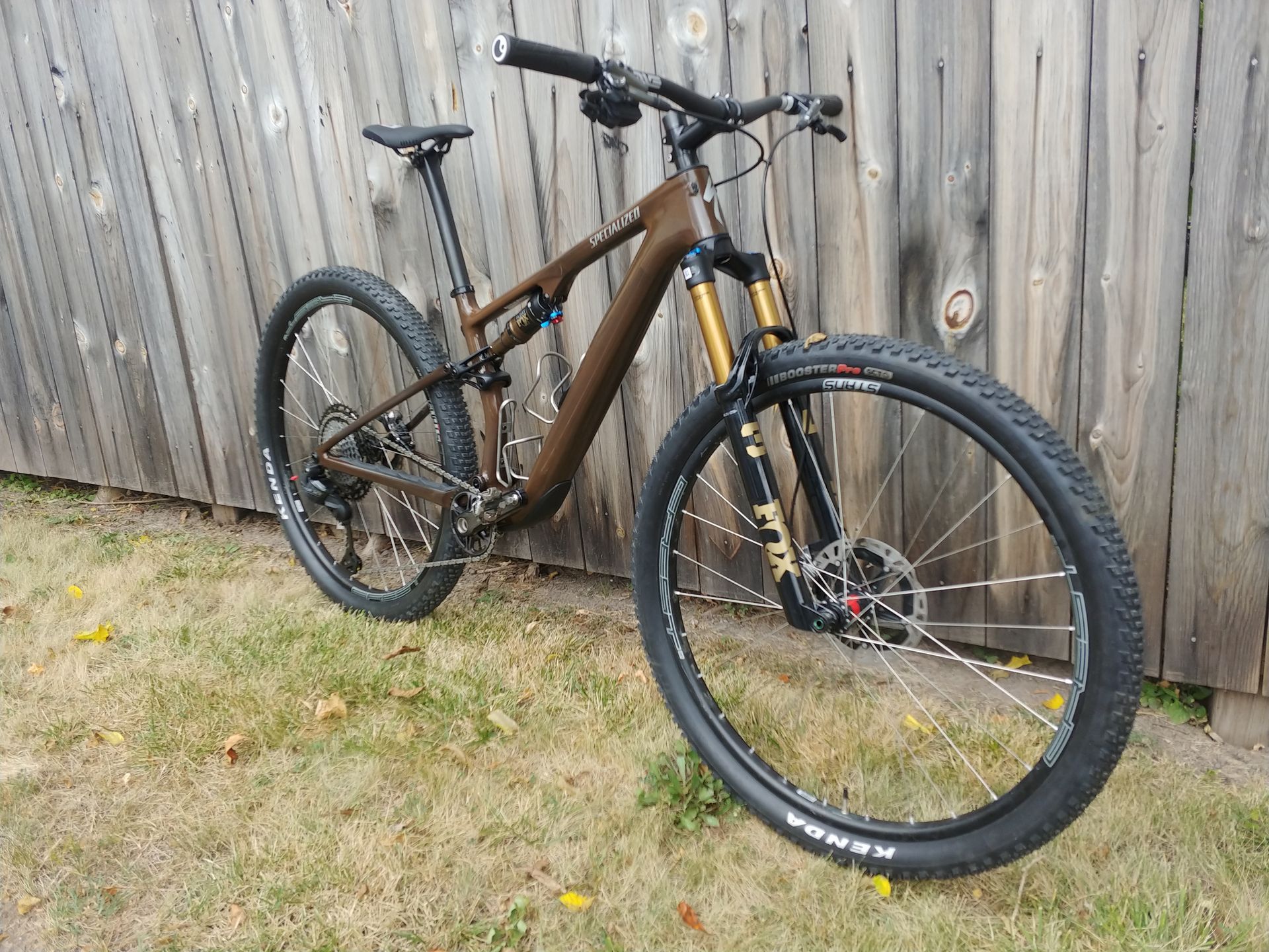 Mountain bike leaning against a wooden fence on grass, with black wheels and a brown frame.