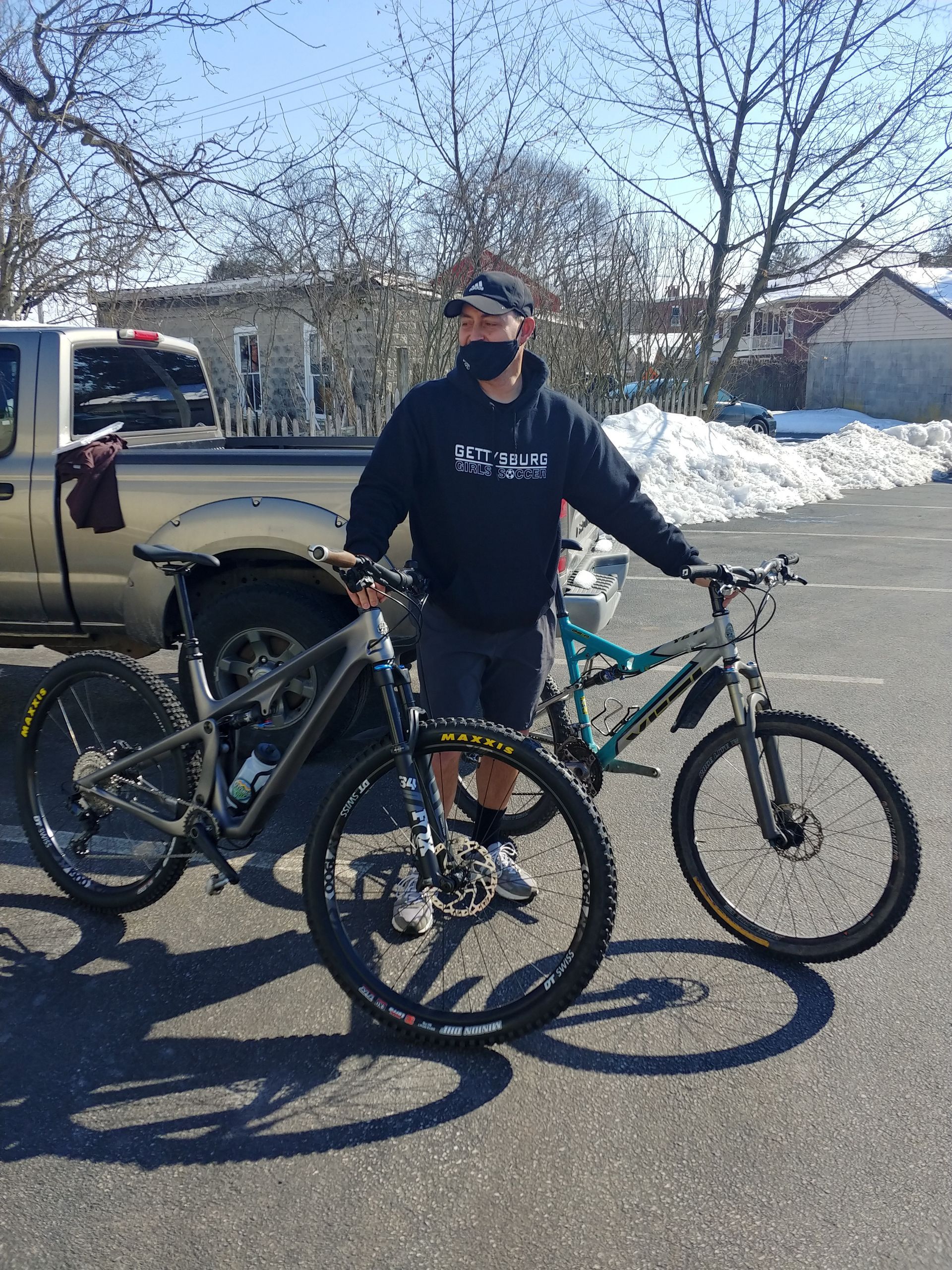 Person standing with two bicycles in a snowy parking lot beside a truck, wearing a helmet and face covering.