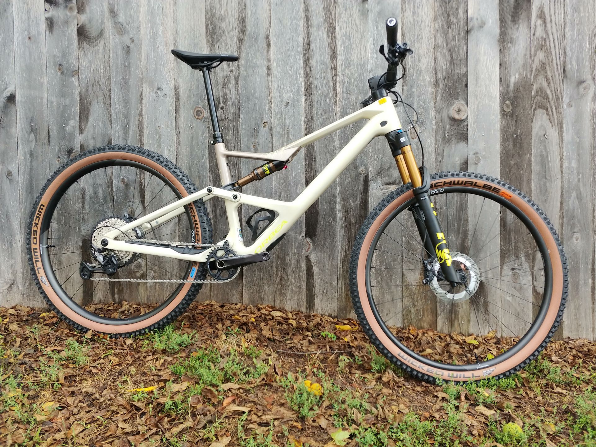 White full-suspension mountain bike leaning against a weathered wooden fence outdoors, with muddy tires.