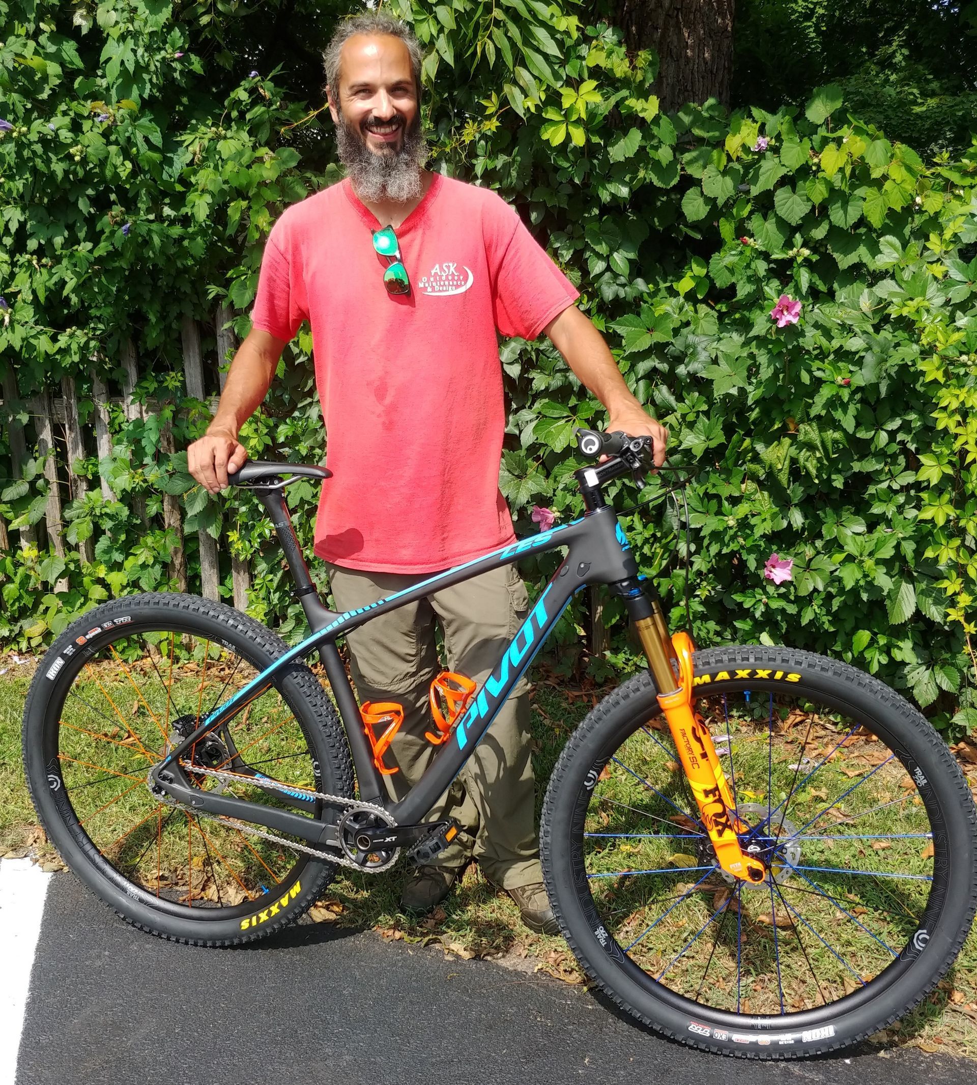 Man standing beside a black and orange mountain bike on a road in front of leafy greenery