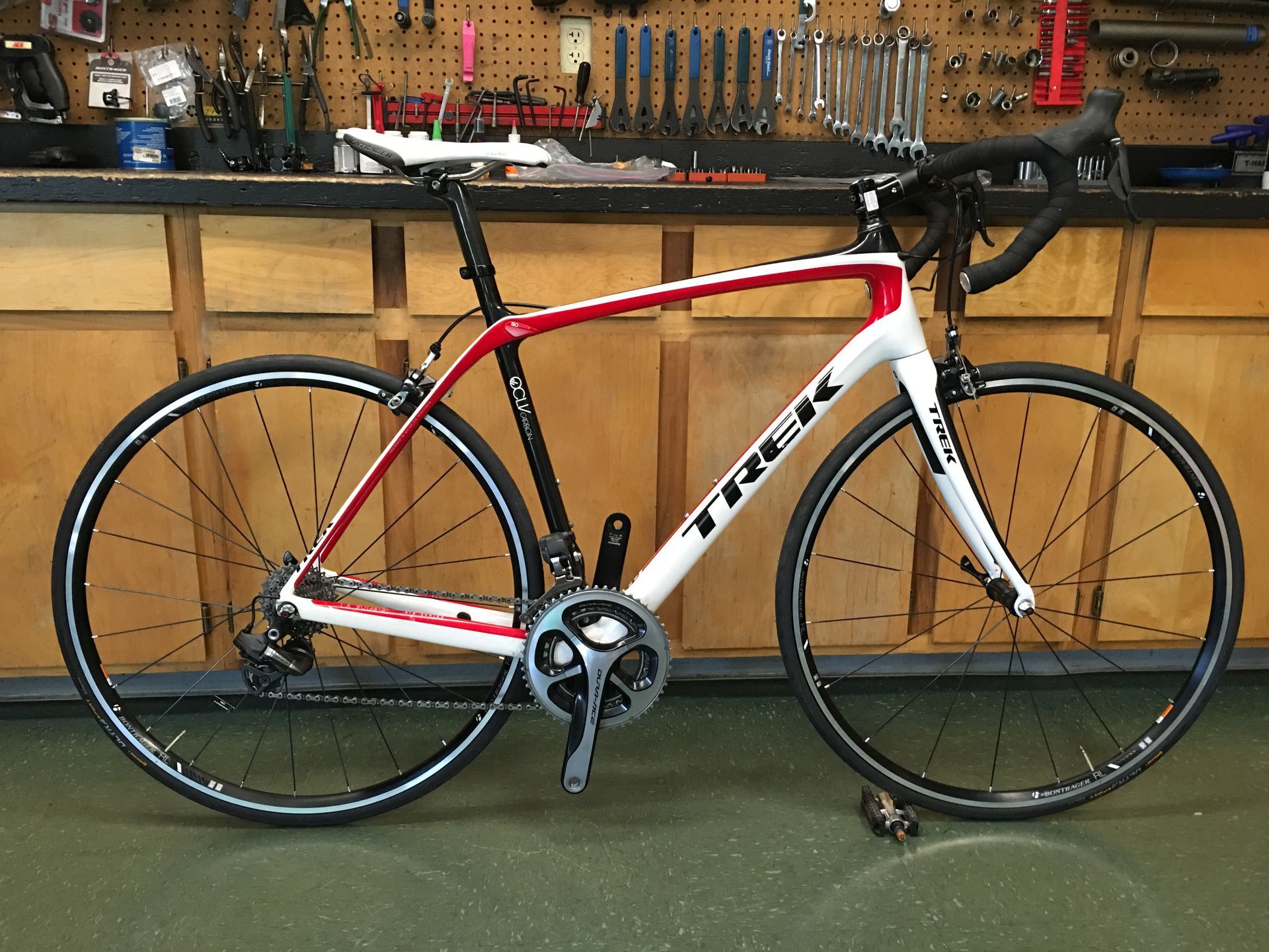 Red and white Trek road bike on a stand in a workshop with tools on the wall.