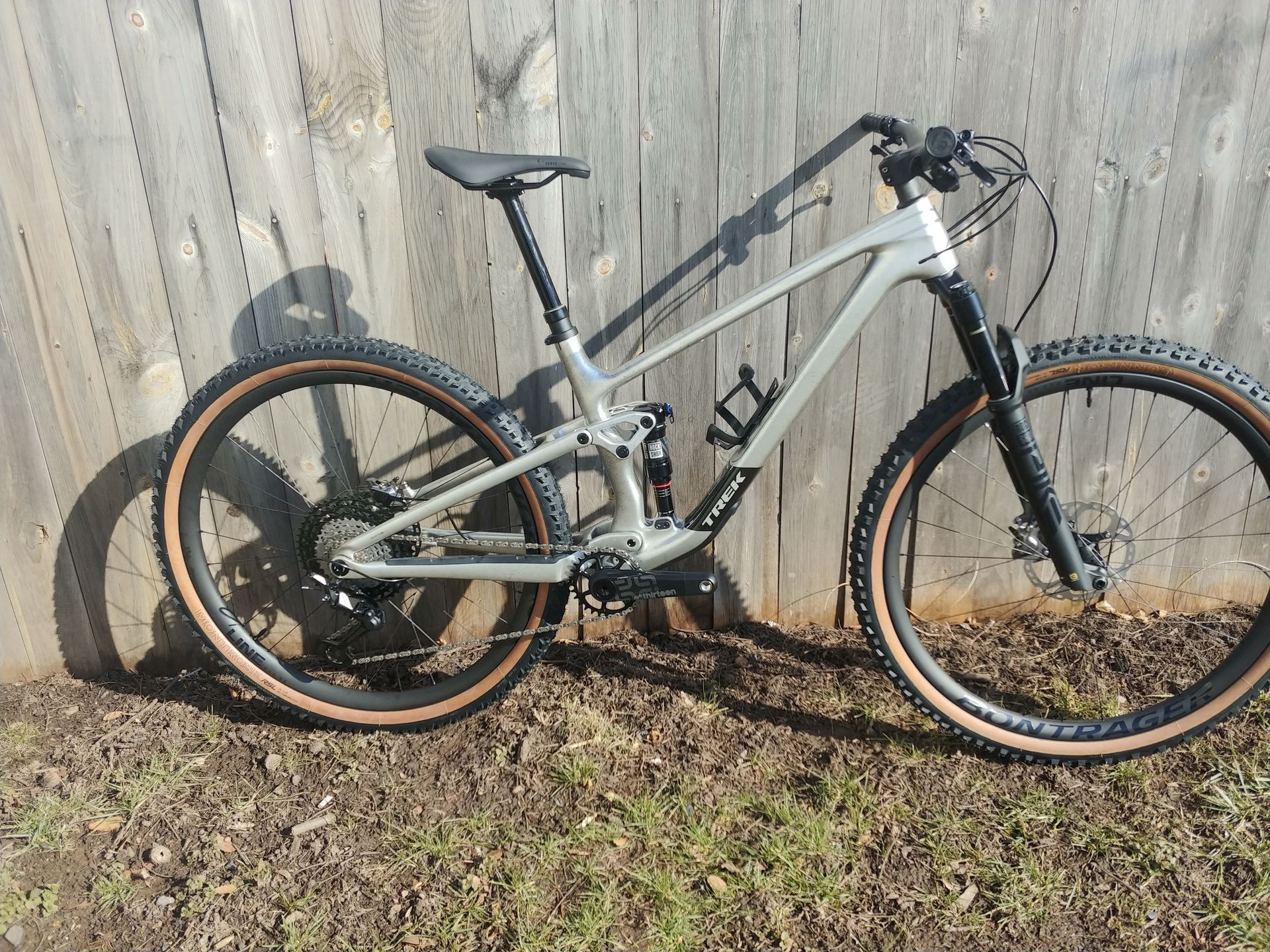 Mountain bike with silver frame and black suspension leaning against a wooden fence outdoors.
