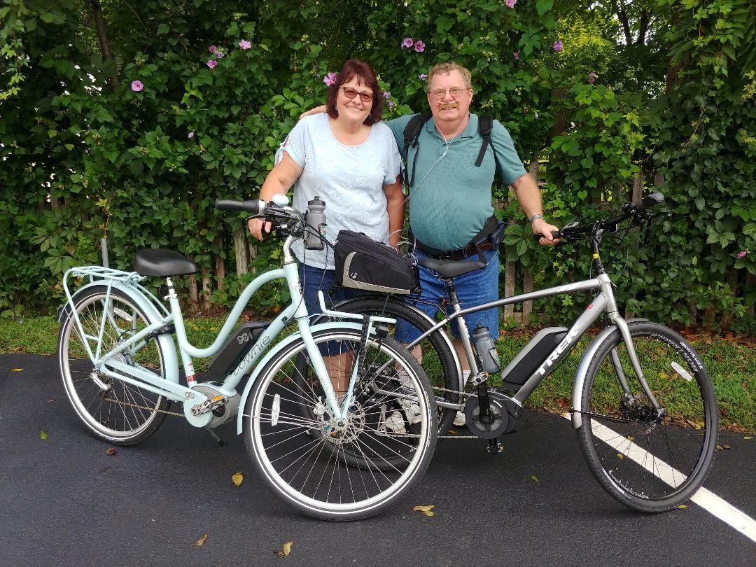 Two people standing with bicycles on a paved path beside dense green bushes