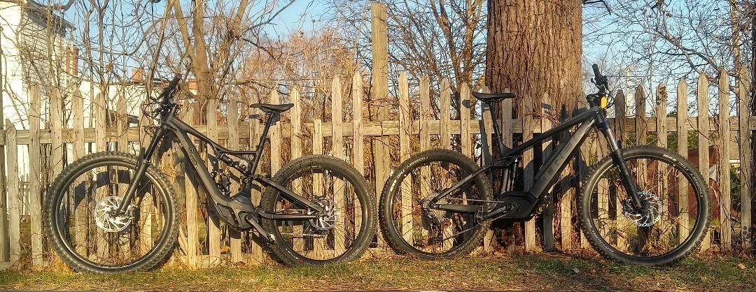 Two black mountain bikes leaning against a wooden fence in a leafless yard.
