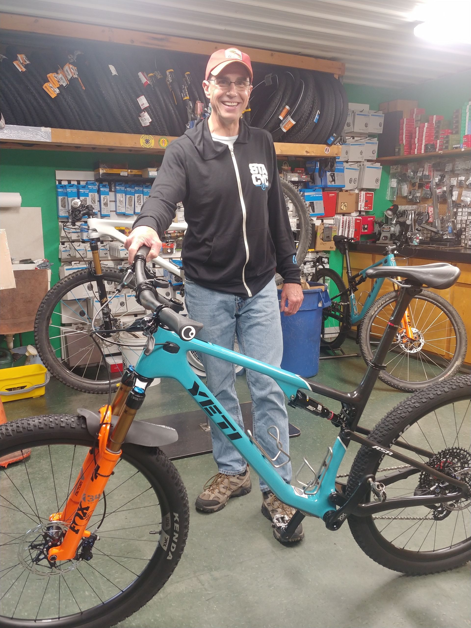 Person stands in a bike shop holding a turquoise mountain bike beside an orange bike.