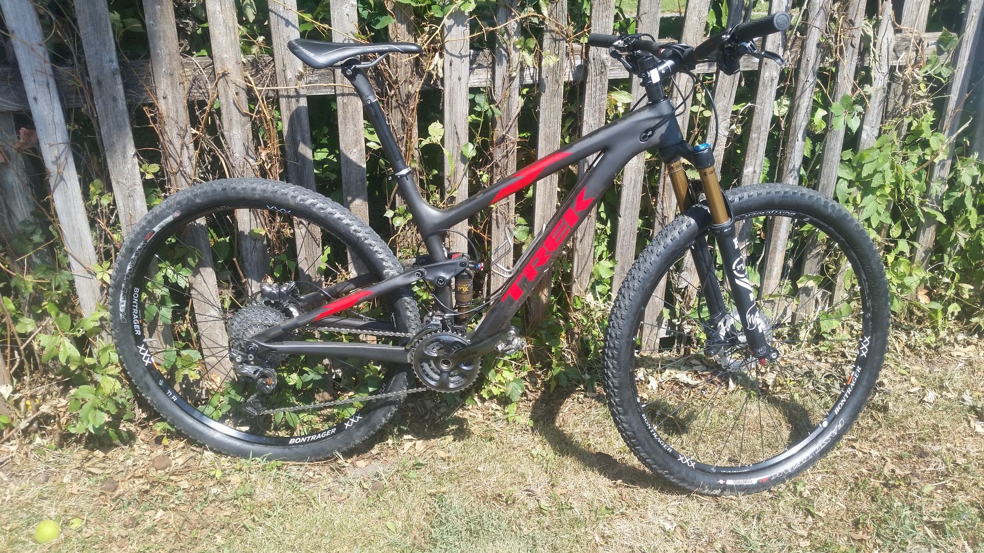 Red and black mountain bike leaning against a wooden fence in a grassy yard.