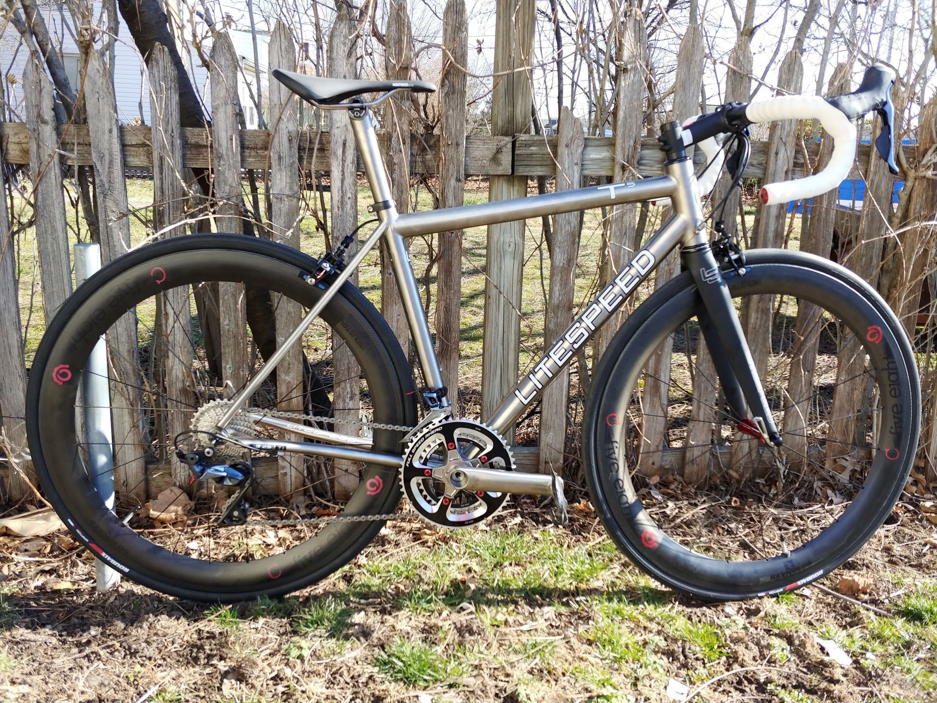 Silver road bike leaning against trees in a wooded area, with black tires and white handlebars.