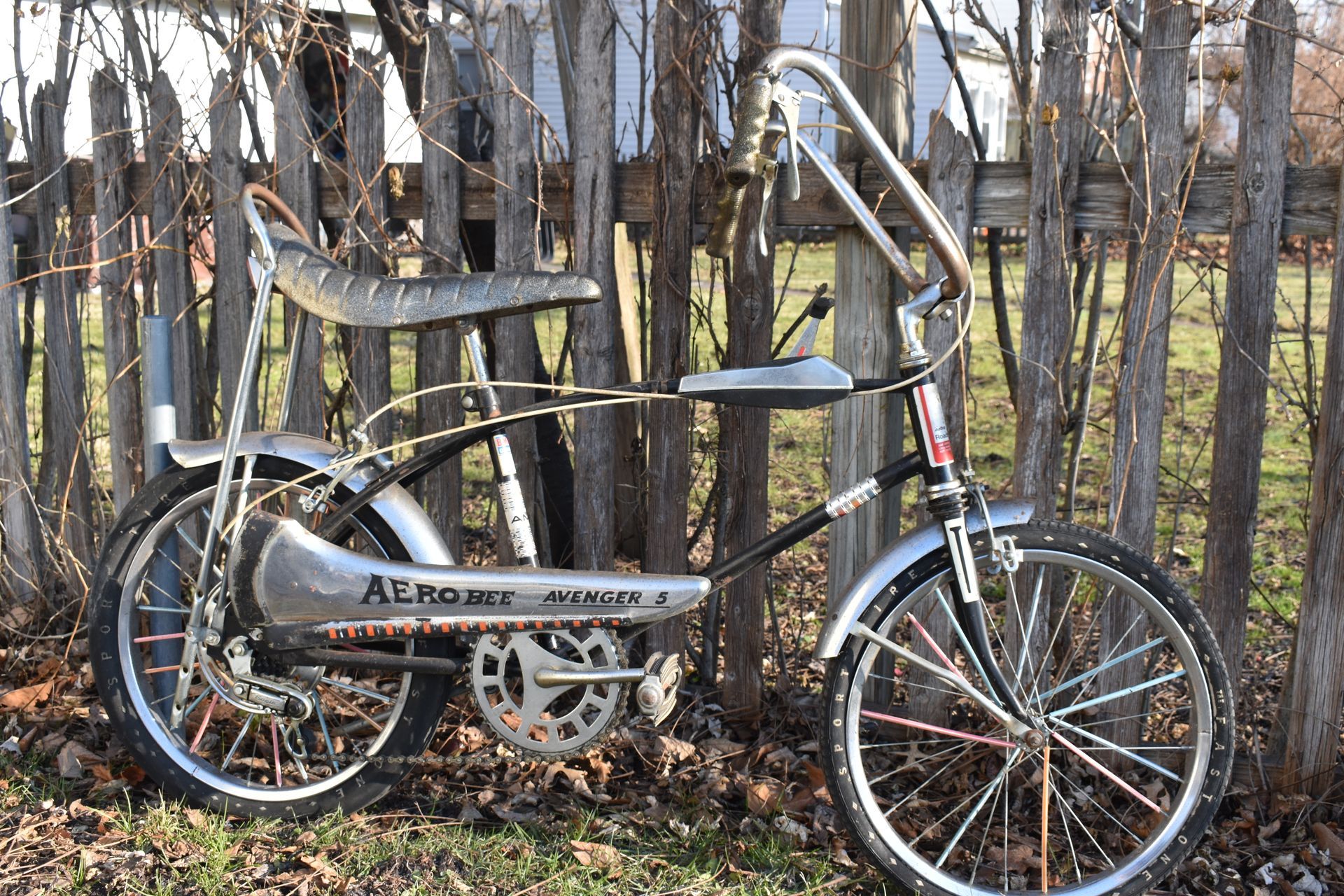 Silver recumbent bicycle parked by a wooden fence in a leafless wooded area