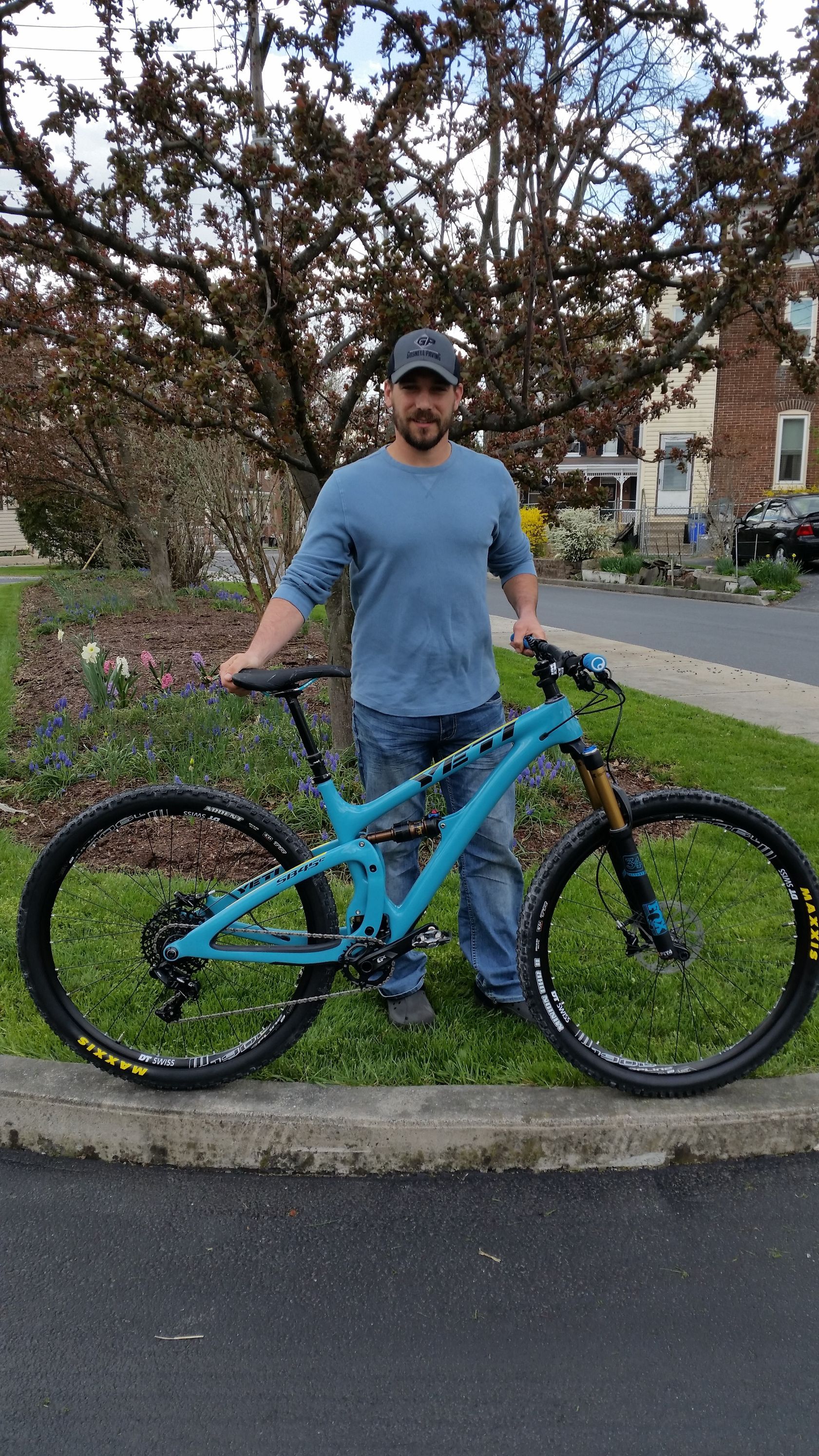 Man standing beside a blue mountain bike on a sidewalk near a grassy curb and trees
