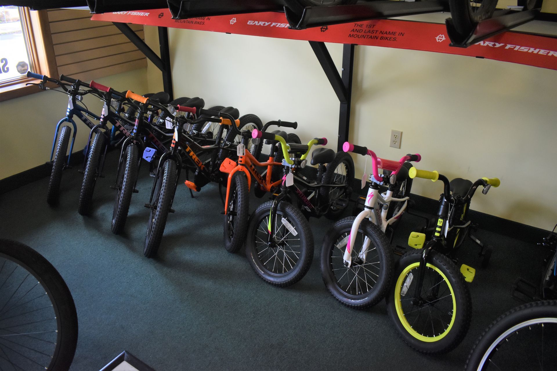Row of bicycles and children’s bikes lined up indoors under a shelf, mostly black with one pink and one yellow bike.