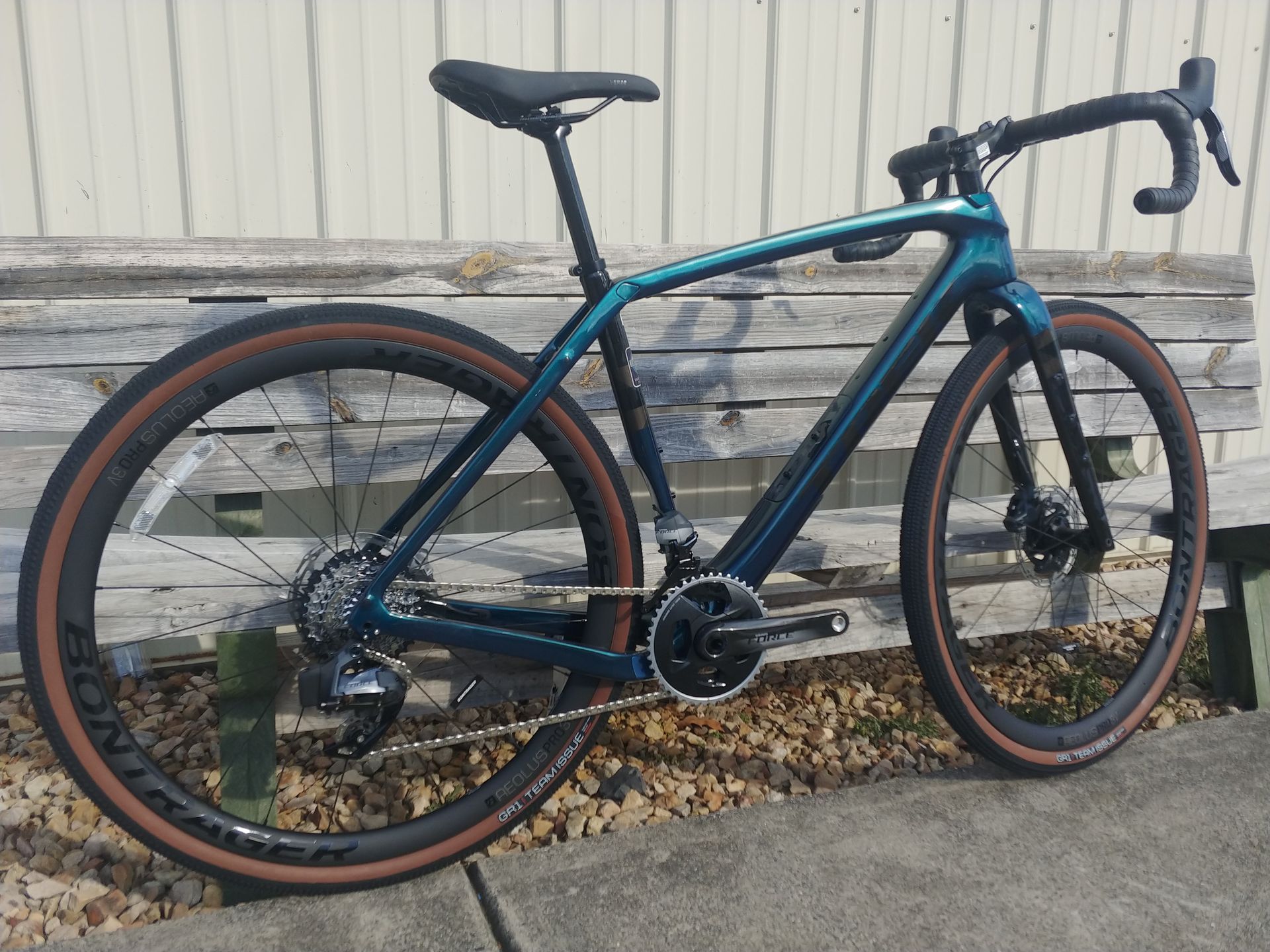 Black and teal road bike leaning against a weathered wooden wall with gravel below