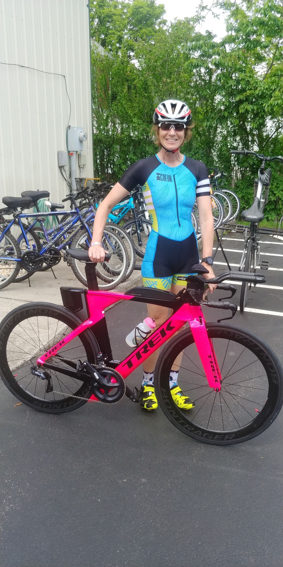 Cyclist in blue jersey stands beside a pink road bike outdoors near several parked bicycles.