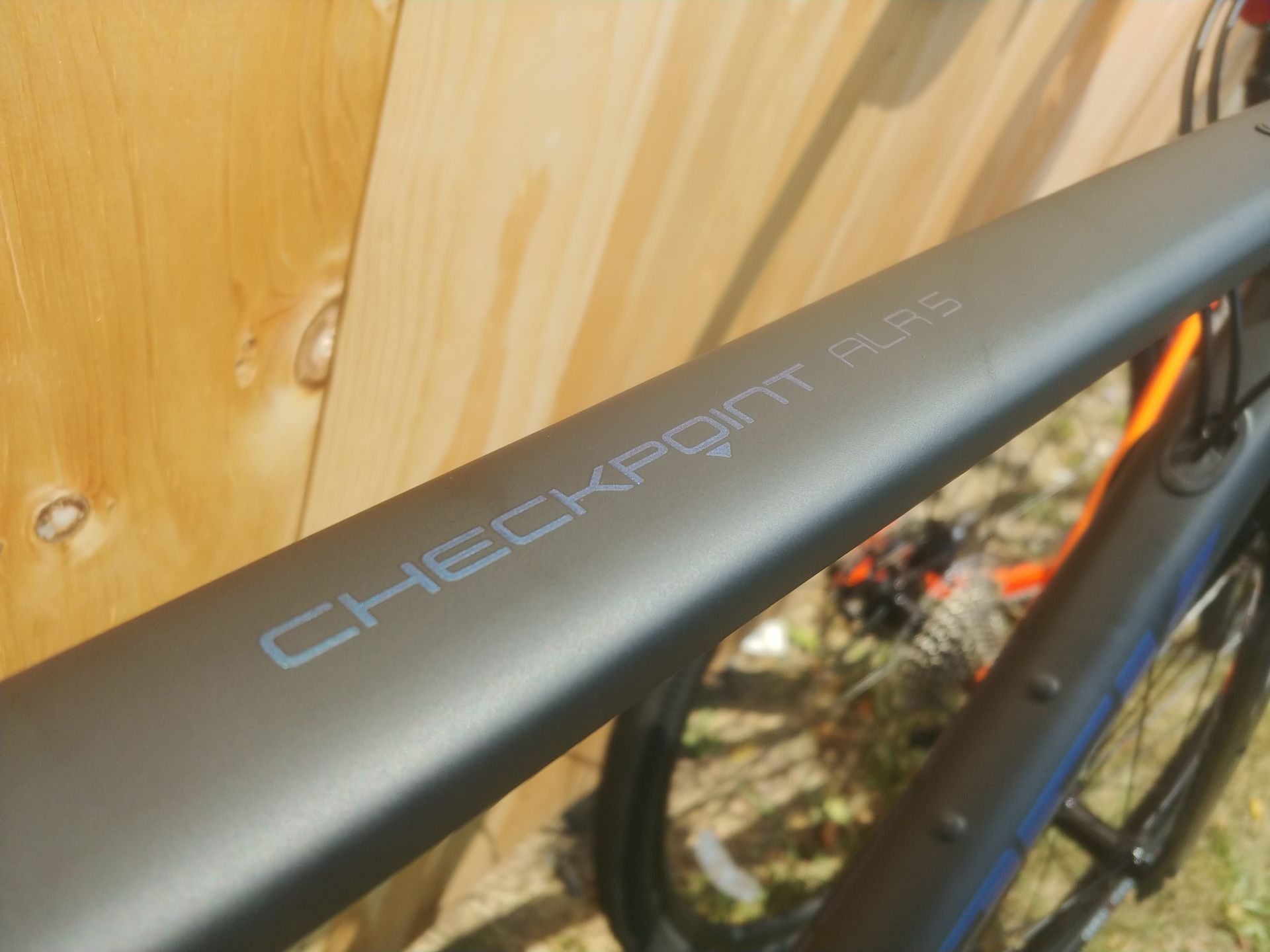 Close-up of a matte black Checkpoint bike frame with blue lettering near a wooden wall.