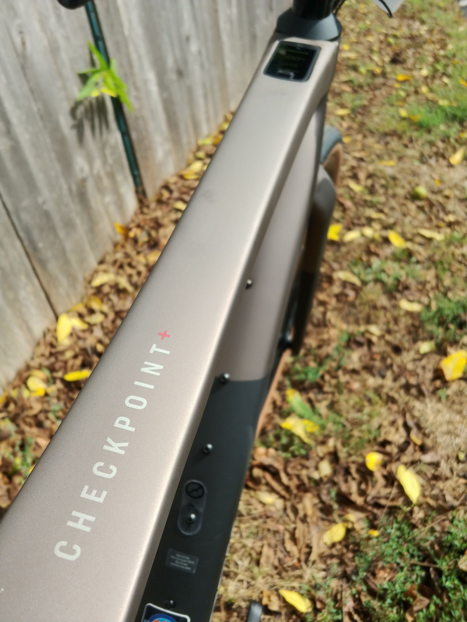 Close-up of a black electric scooter deck on wet leaves beside a wooden fence