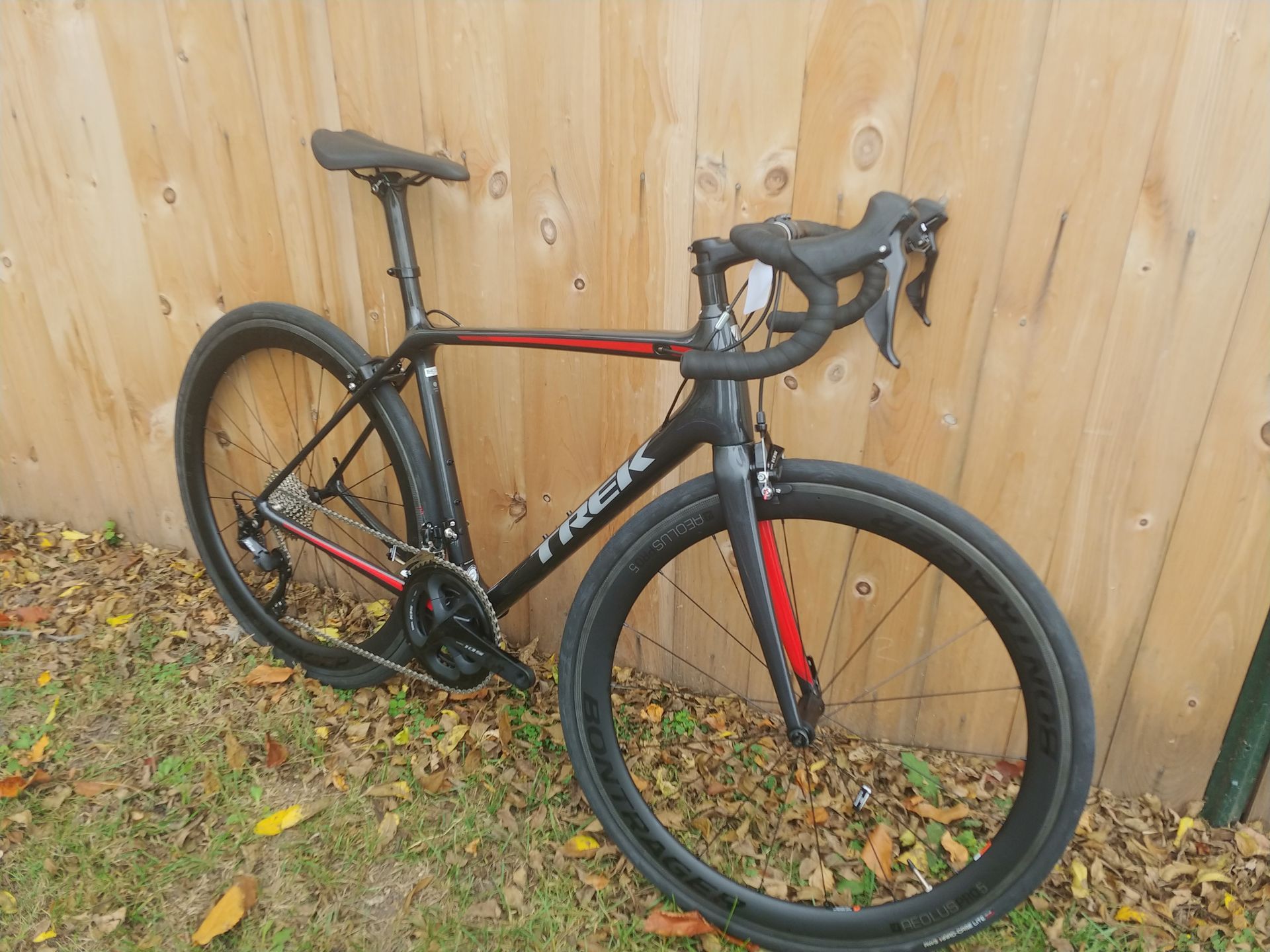 Black and red road bike leaned against a wooden fence on leaf-covered ground.