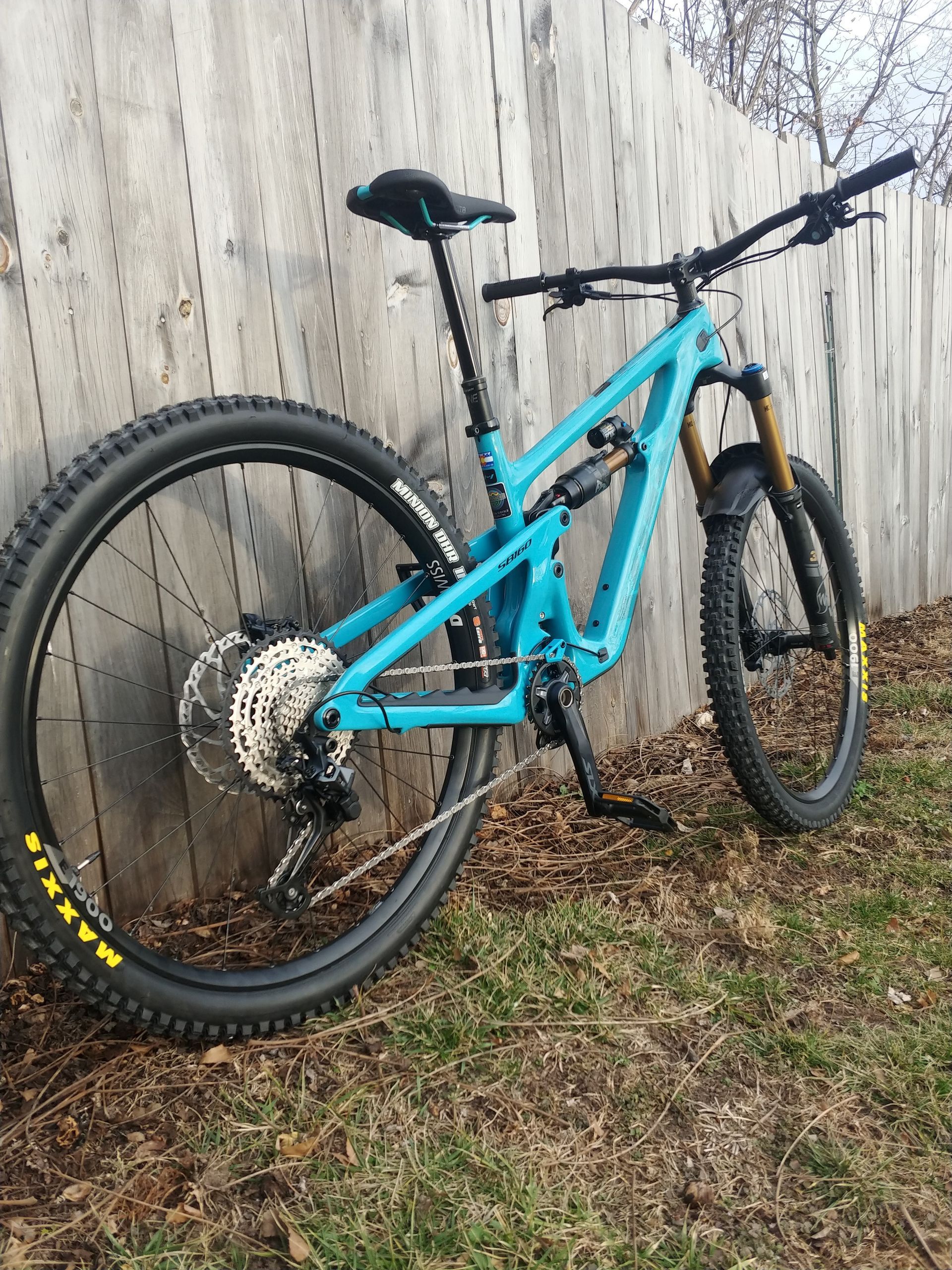 Teal mountain bike leaning against a wooden fence on grass, with muddy tires.