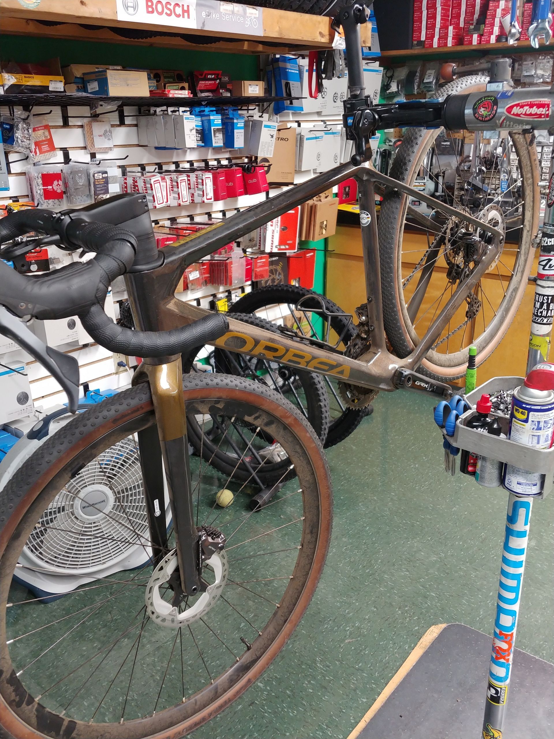 Mud-covered bicycle with drop handlebars in a shop aisle, beside other bikes and shelving.