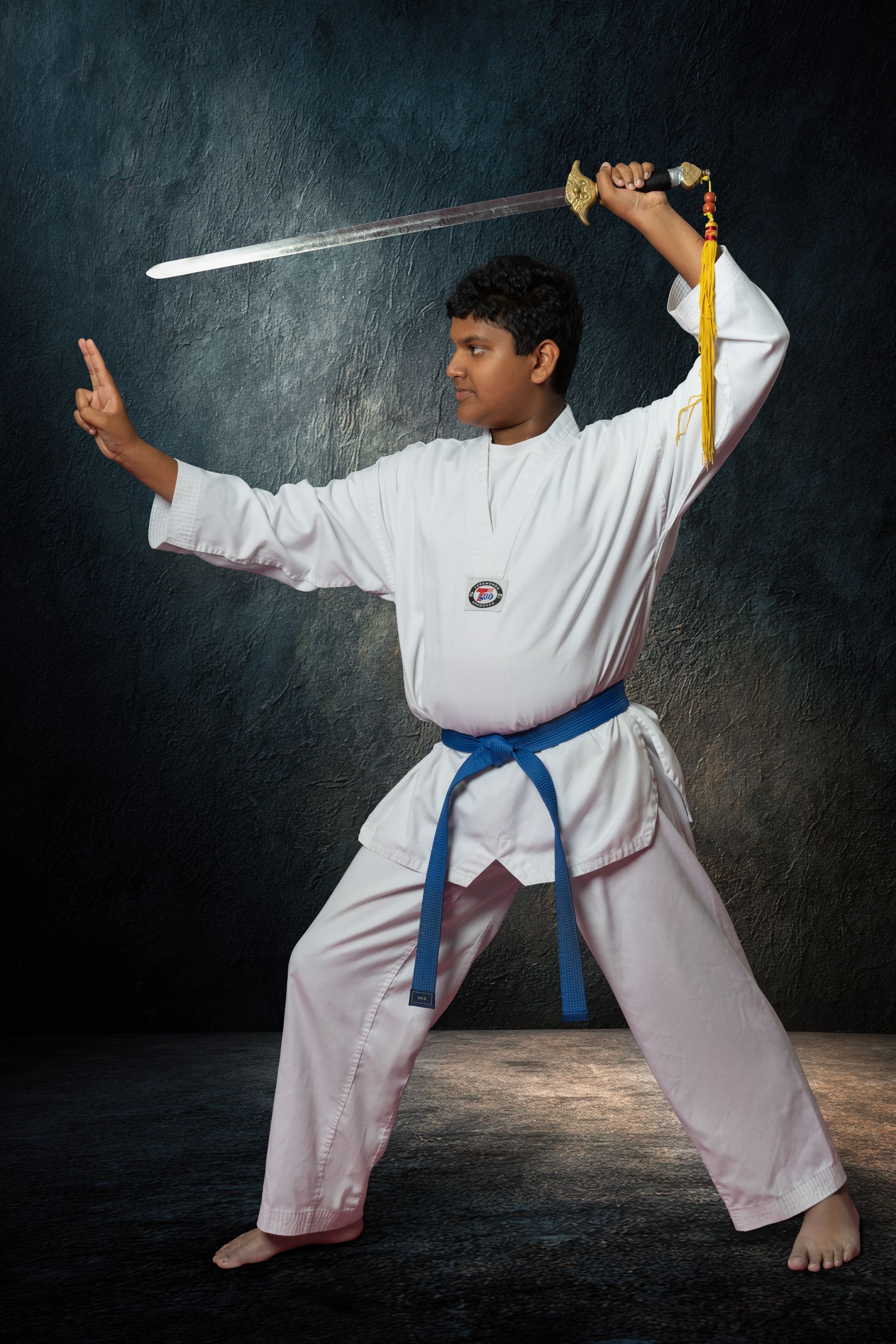 Martial artists in uniforms stand in a circle on an outdoor stage as a person in the center jumps to break wooden boards.