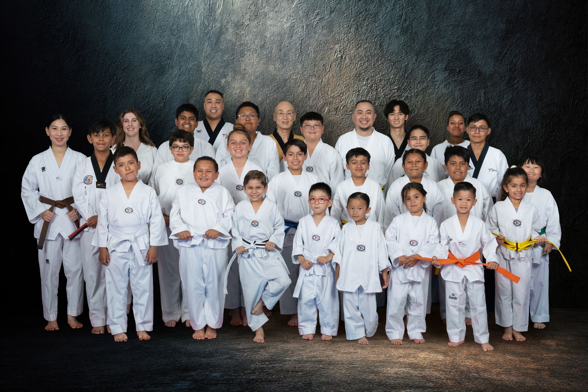 A group of taekwondo students and instructors pose with a branded academy flag in a training studio.