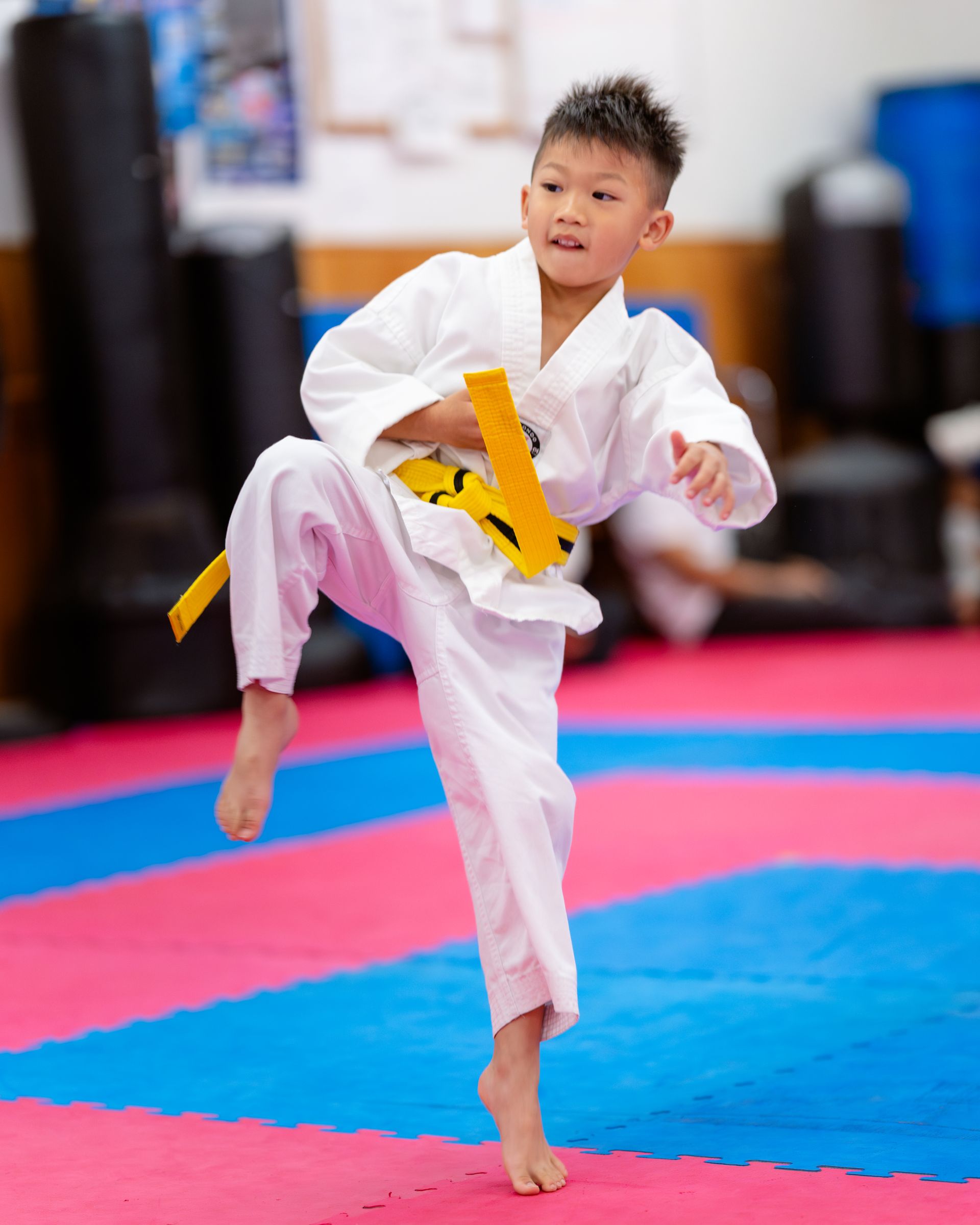 Children in martial arts uniforms and protective vests walking in an indoor event space with a Taekwondo banner.