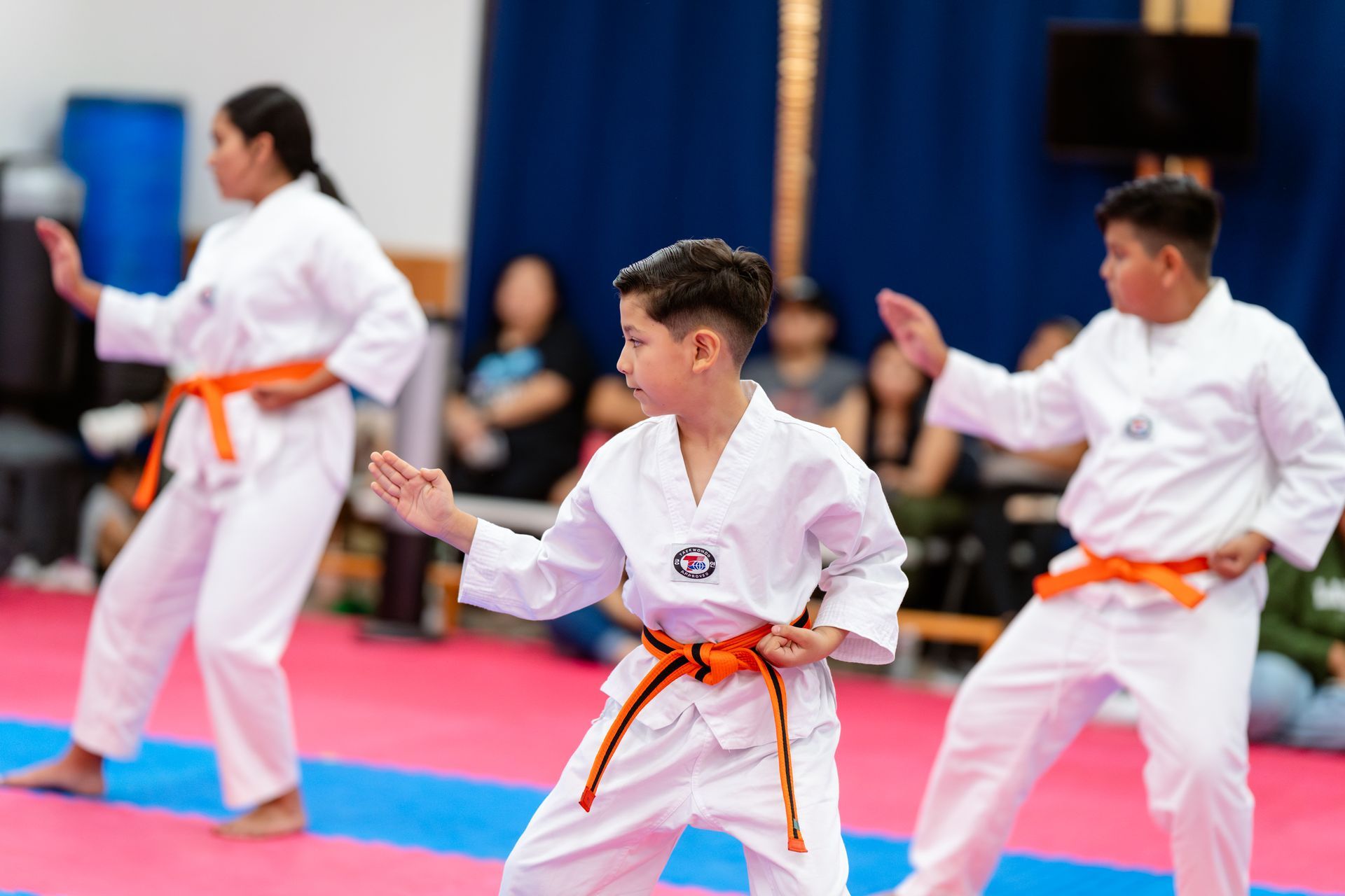 Children in martial arts uniforms stand on a red and blue padded mat in a large indoor competition venue.
