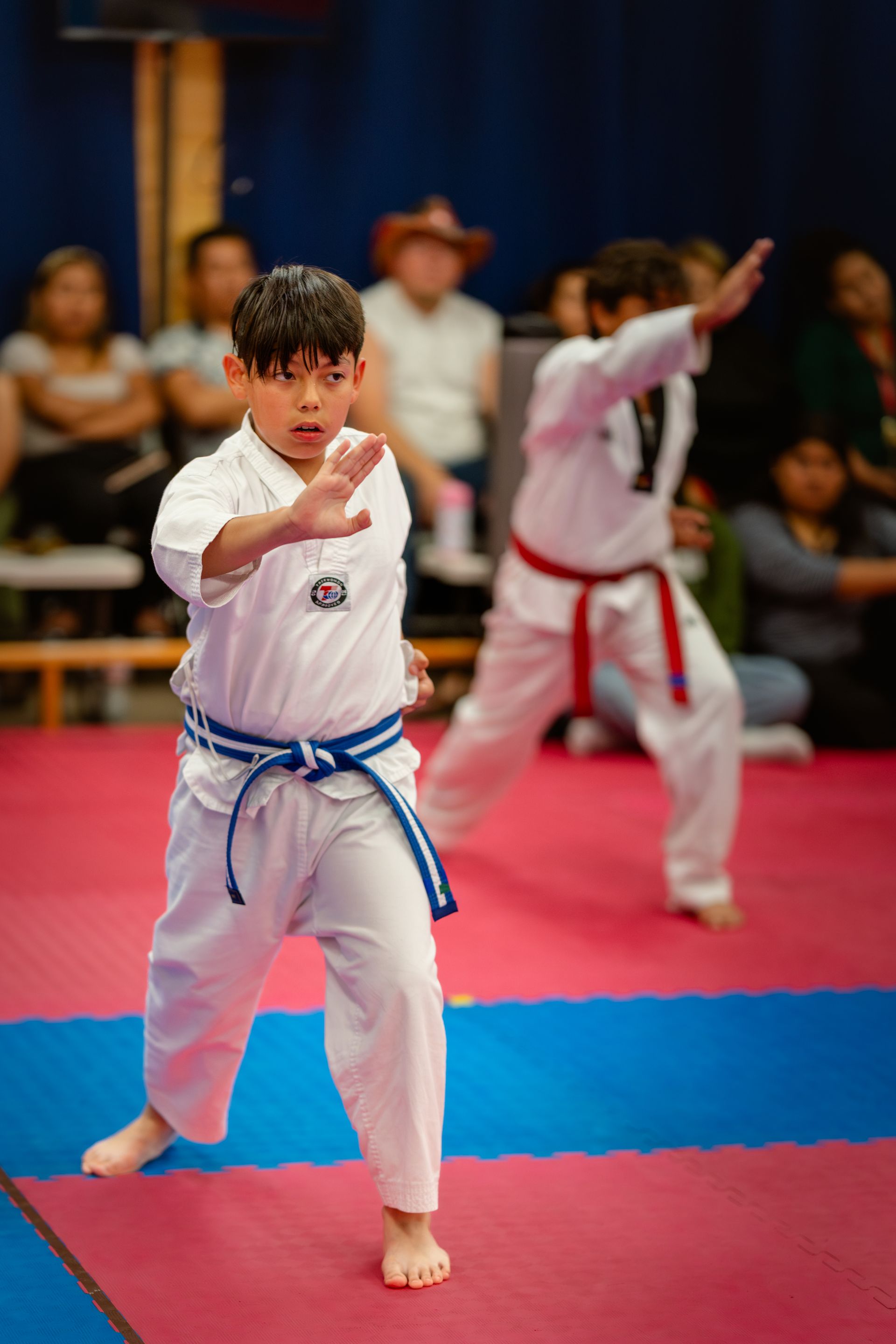 People in taekwondo uniforms practice sparring and drills in a gym with red and blue mats.