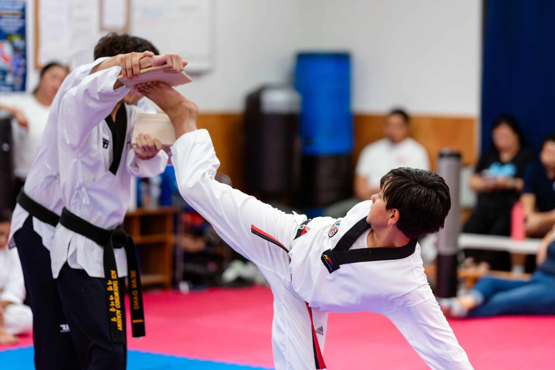 A sparring match in a taekwondo studio with participants wearing white uniforms and protective head and chest gear.