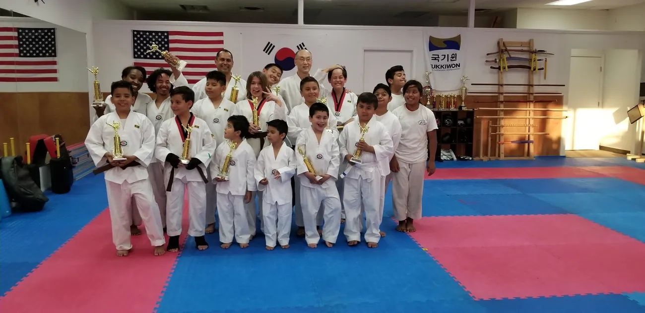 A group of martial arts students in white uniforms standing in a training studio with flags and trophies in the back.