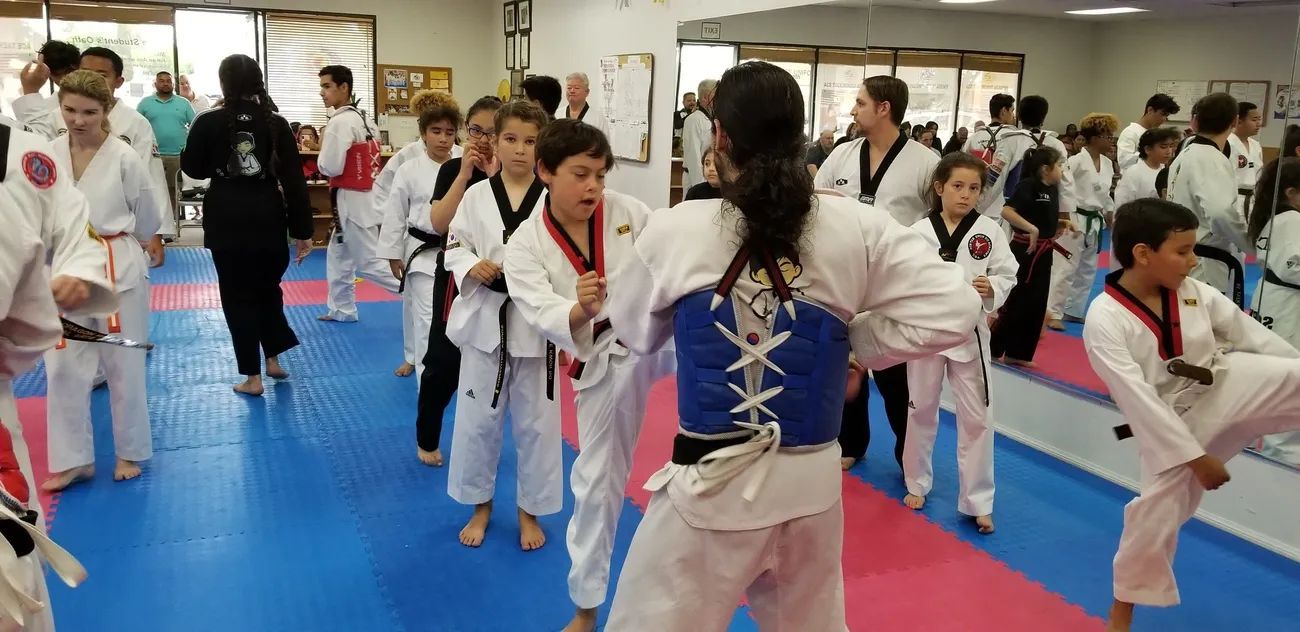 Students in white martial arts uniforms with black belts practice taekwondo in a training studio with mats.