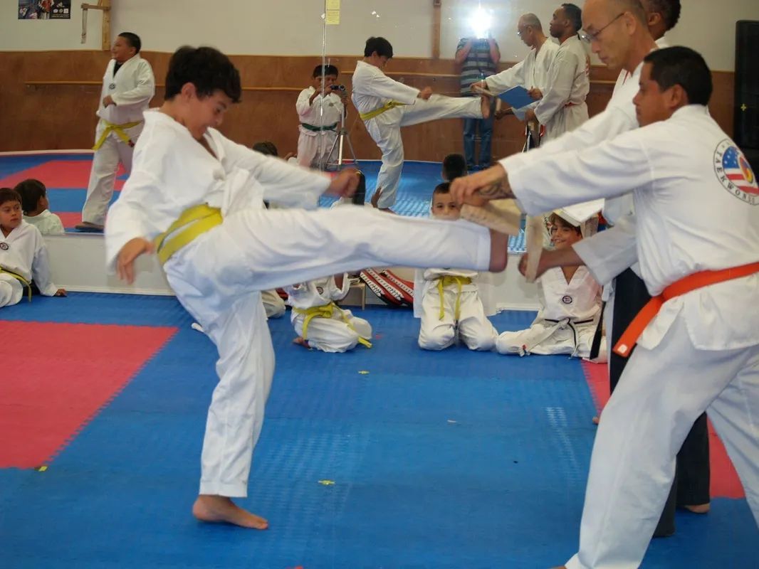A student in a white uniform with a yellow belt kicks a board held by an instructor in an orange belt in a martial arts gym.