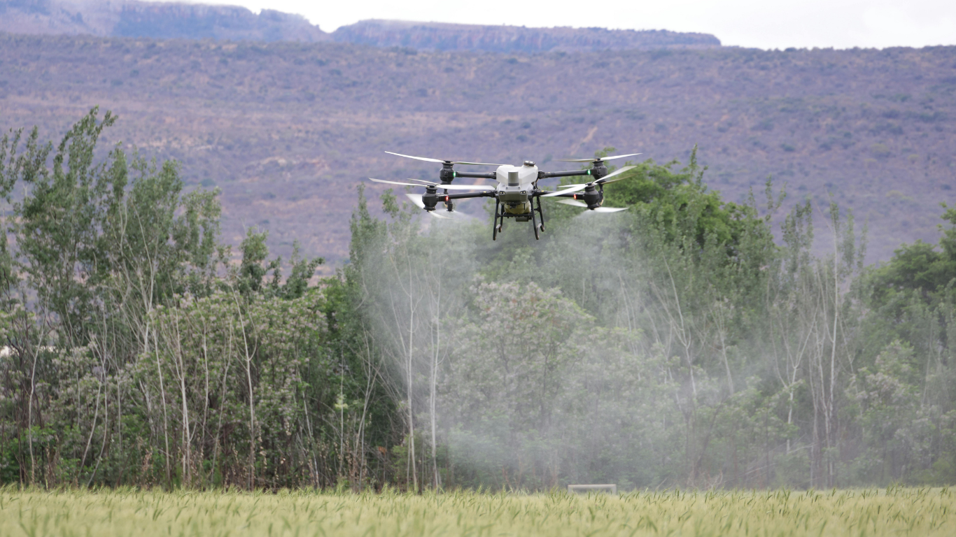 An agricultural drone flies over a green field, spraying a mist of liquid over the crops against a backdrop of hills.