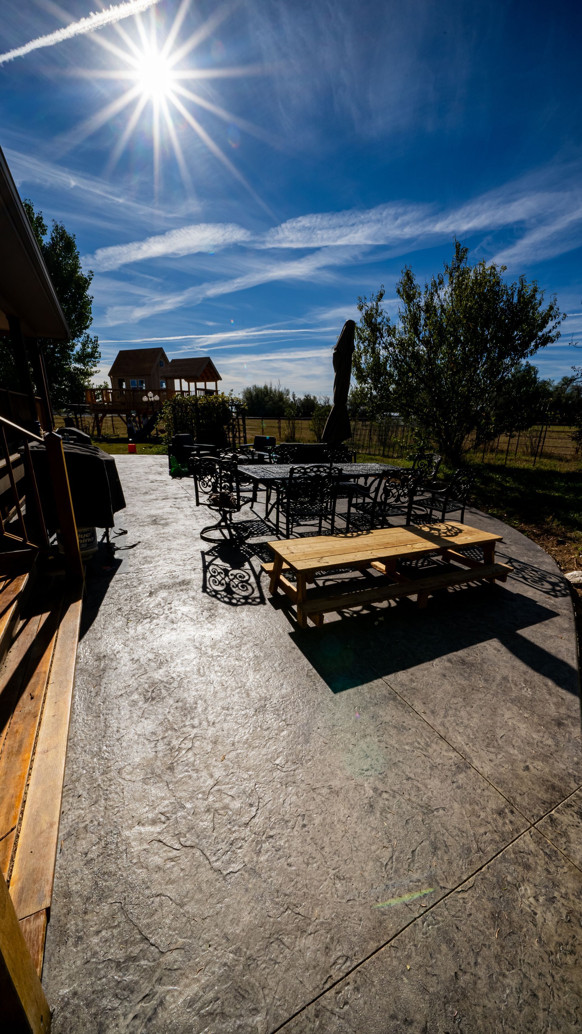 Sunlit patio with benches, picnic tables, and trees under a bright blue sky with the sun overhead