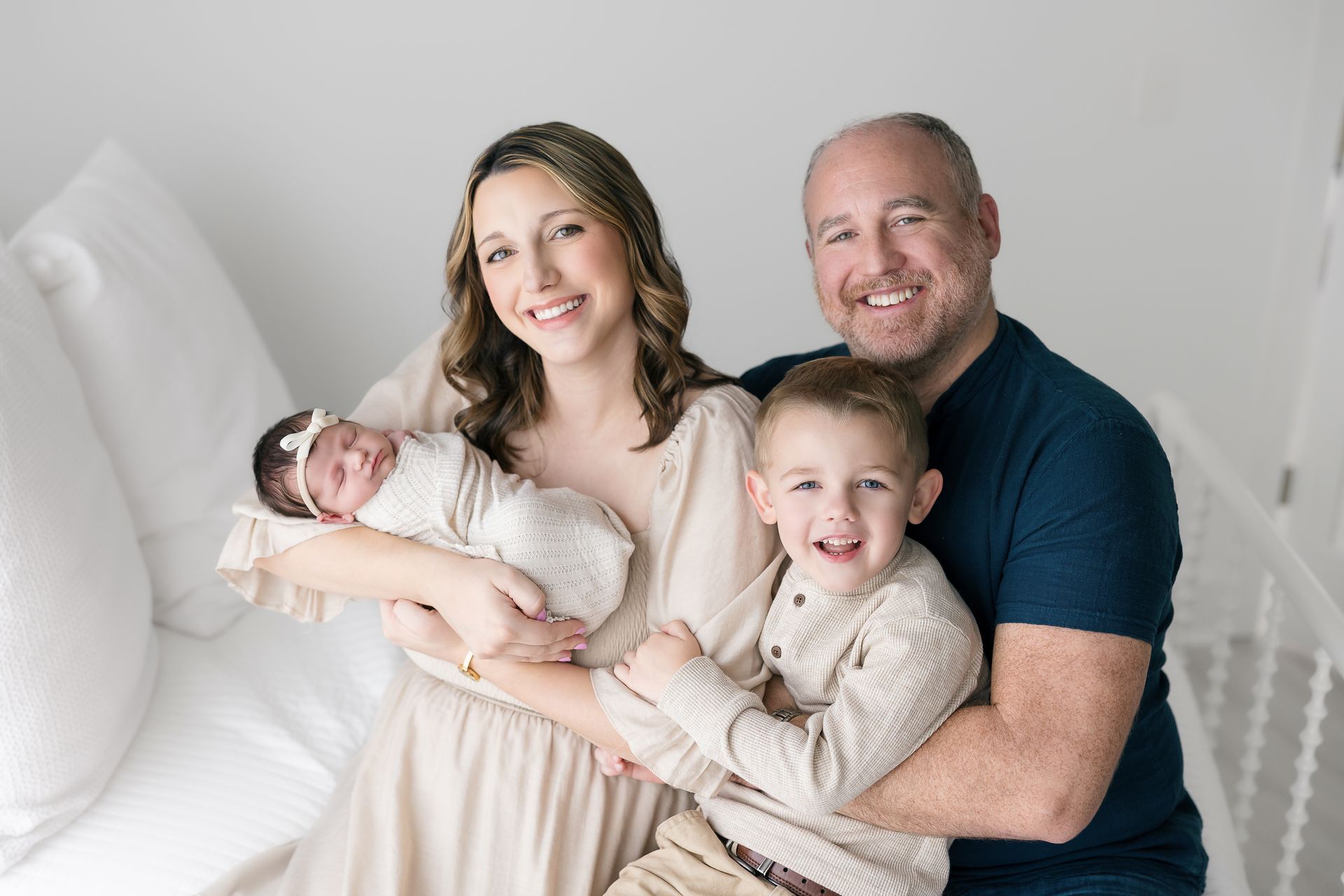 Family portrait on white stairs: smiling adults holding a baby and toddler in light clothing