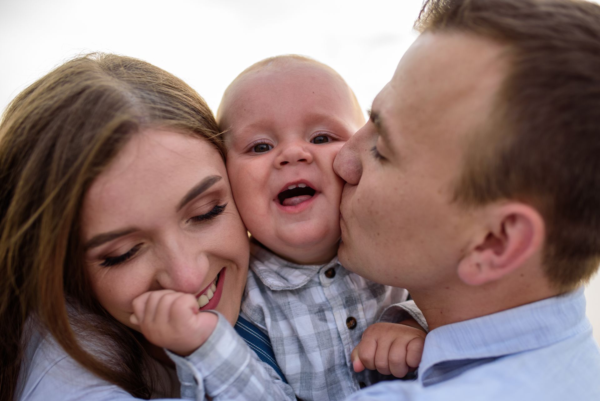 A smiling baby is kissed on both cheeks by two adults against a white background