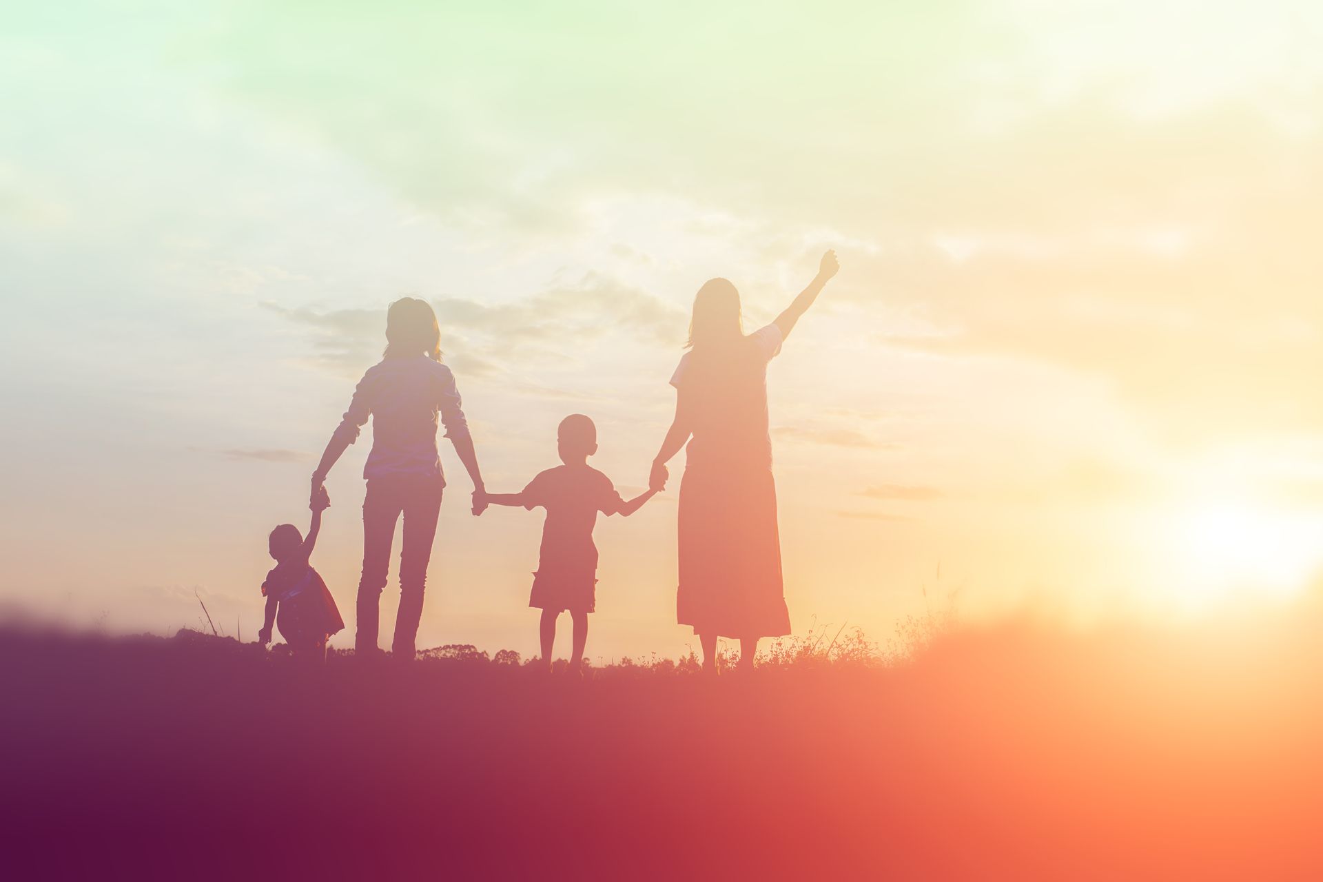 Silhouetted family of four holding hands on a hill at sunset with warm orange light