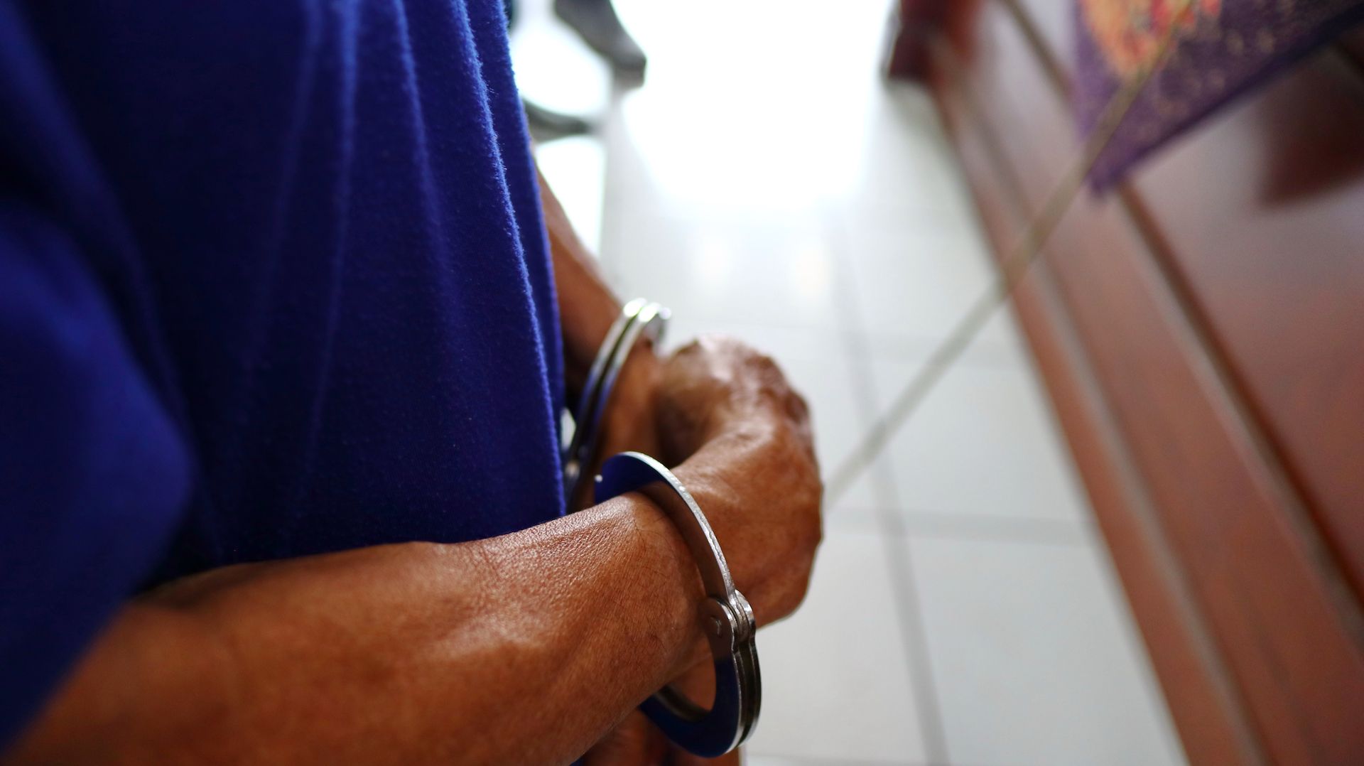 Hands cuffed behind back in a tiled hallway, with a person in a blue shirt visible at left