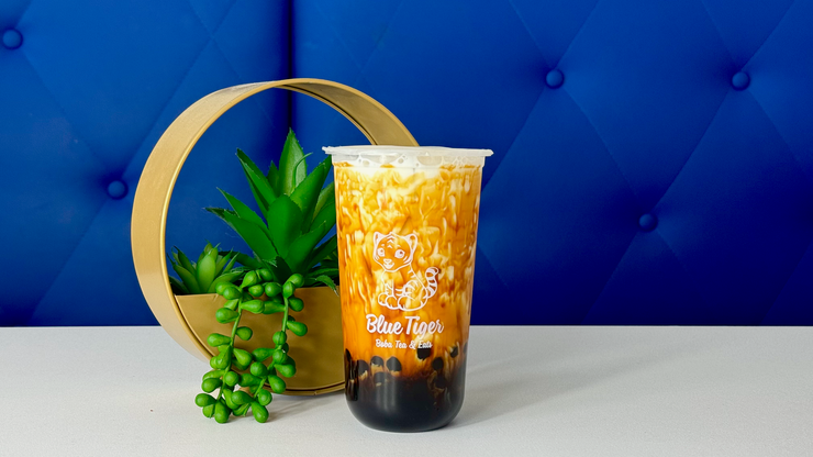 Red iced drink in a clear cup beside a green plant and gold hoop on a white surface