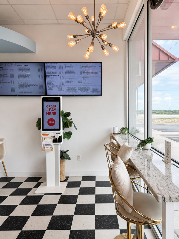Bright café interior with black-and-white checkered floor, menu screens, and window seating.
